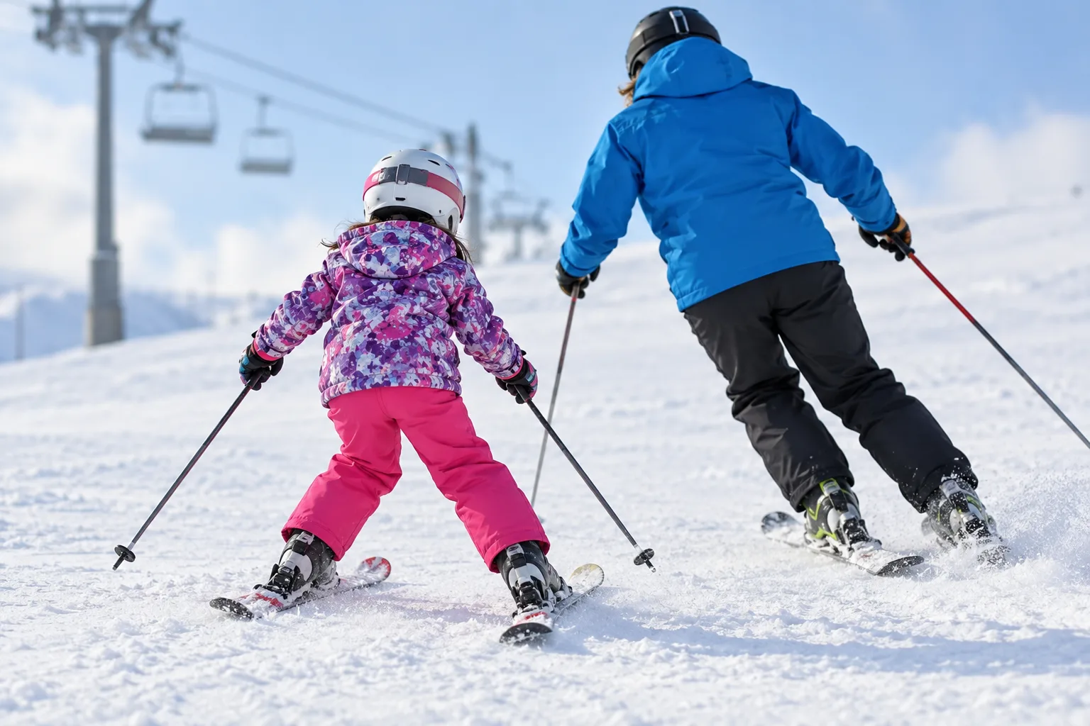 Children in colorful ski gear practice turns with an instructor on a snowy beginner slope beside a lift.